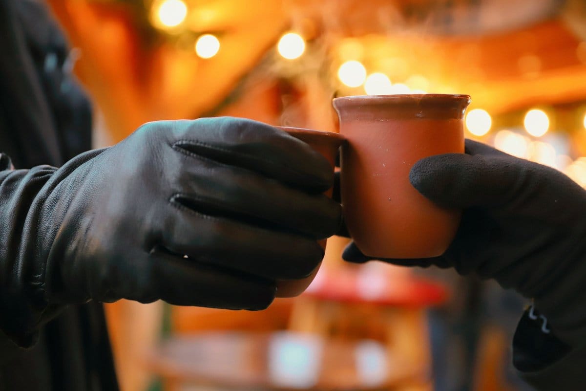 Zwei Hände in Handschuhen stoßen mit Glühweintassen vor warmen Lichterketten an - Symbolbild für den Soester Weihnachtsmarkt